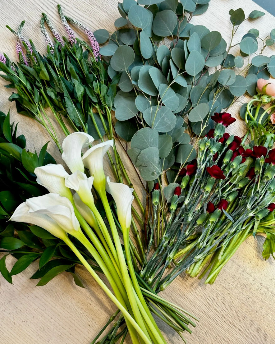 Flat-lay of mixed floral stems on a light wood table: white calla lilies, silver-dollar eucalyptus, burgundy spray carnations, pink veronica spikes and lush greenery ready for bouquet arranging.