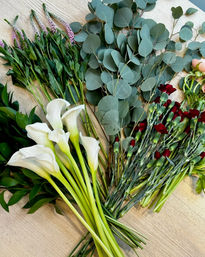 Flat-lay of mixed floral stems on a light wood table: white calla lilies, silver-dollar eucalyptus, burgundy spray carnations, pink veronica spikes and lush greenery ready for bouquet arranging.