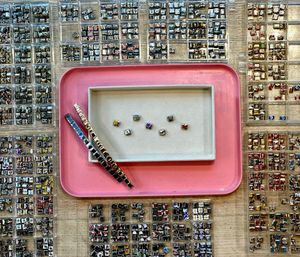 Top-down view of a jewelry display with hundreds of tiny square metal charm links and colorful inserts sorted in clear plastic trays around a pink tray and beige dish holding a few loose sparkly charms and two bracelet strips.
