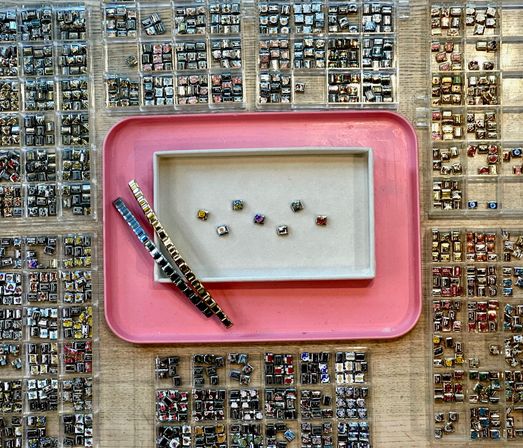 Top-down view of a jewelry display with hundreds of tiny square metal charm links and colorful inserts sorted in clear plastic trays around a pink tray and beige dish holding a few loose sparkly charms and two bracelet strips.