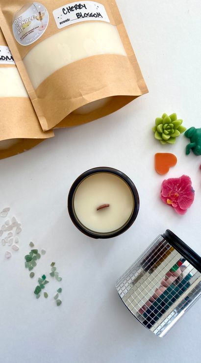 Flat-lay of DIY candle-making supplies on white background: two kraft pouches of scented wax (cherry and cactus blossom), an amber jar soy candle with wooden wick, colorful shaped wax melts (succulent, frog, heart, butterfly, flower), scattered sea-glass chips and a small mirrored jar.