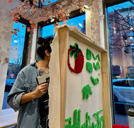 Person working on punch-needle textile art: framed canvas with a red strawberry and green tufted shapes in a cozy craft studio window display with hanging cherry blossoms and city street outside.