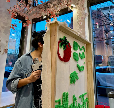 Person working on punch-needle textile art: framed canvas with a red strawberry and green tufted shapes in a cozy craft studio window display with hanging cherry blossoms and city street outside.