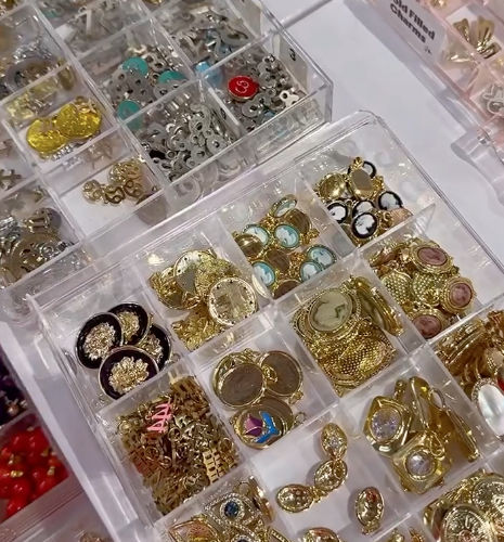 Clear plastic organizer tray of gold-tone charms, cameo pendants, decorative buttons and metal findings — jewelry supplies at a craft market or bead shop display.