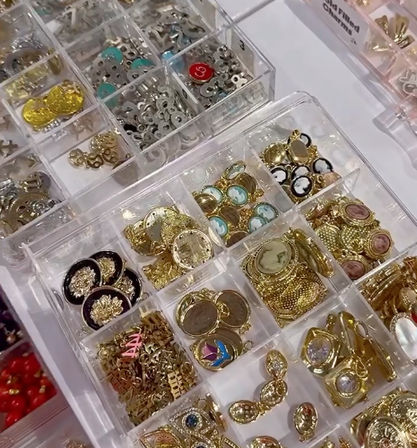 Clear plastic organizer tray of gold-tone charms, cameo pendants, decorative buttons and metal findings — jewelry supplies at a craft market or bead shop display.