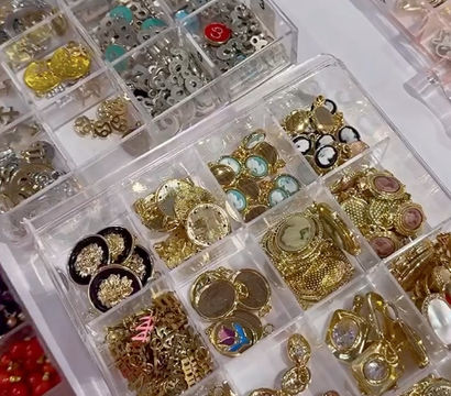 Clear plastic organizer tray of gold-tone charms, cameo pendants, decorative buttons and metal findings — jewelry supplies at a craft market or bead shop display.