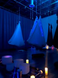 Blue-lit indoor meditation studio with suspended aerial yoga hammocks, glowing candles and crystal singing bowls set up for a relaxing sound bath.