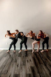 Four women in black activewear lined up in a playful bent-forward yoga pose in a bright indoor fitness studio with wood floors, one woman balancing a small white dog on her lower back.