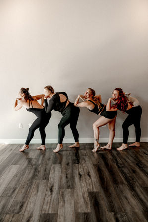 Four women in black activewear lined up in a playful bent-forward yoga pose in a bright indoor fitness studio with wood floors, one woman balancing a small white dog on her lower back.