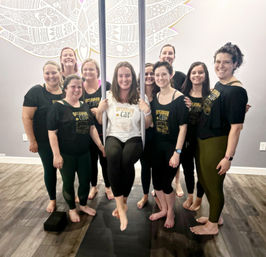 Smiling group of women in an indoor aerial yoga class — one woman seated in an aerial silk hammock while others stand barefoot on mats in a bright studio with a decorative wall mural.