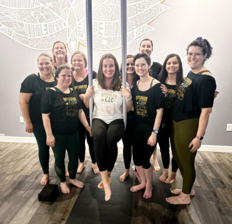 Smiling group of women in an indoor aerial yoga class — one woman seated in an aerial silk hammock while others stand barefoot on mats in a bright studio with a decorative wall mural.