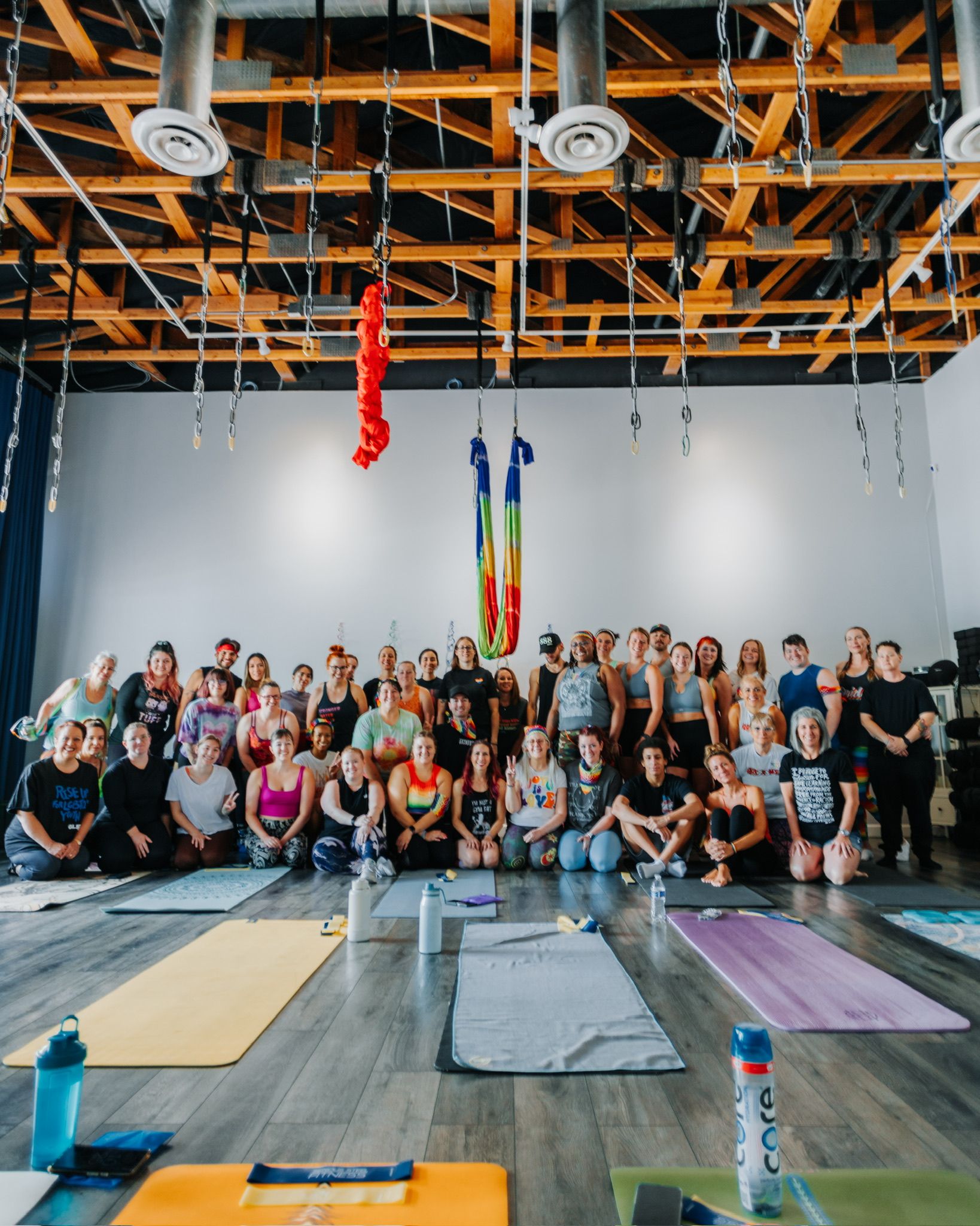 Aerial yoga workshop group in an airy studio with exposed wooden beams, rainbow silks hanging above, rows of yoga mats and water bottles on the hardwood floor