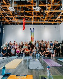 Aerial yoga workshop group in an airy studio with exposed wooden beams, rainbow silks hanging above, rows of yoga mats and water bottles on the hardwood floor