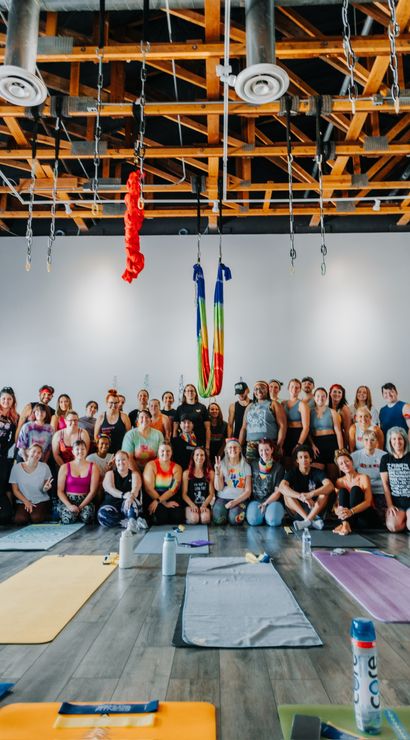 Aerial yoga workshop group in an airy studio with exposed wooden beams, rainbow silks hanging above, rows of yoga mats and water bottles on the hardwood floor