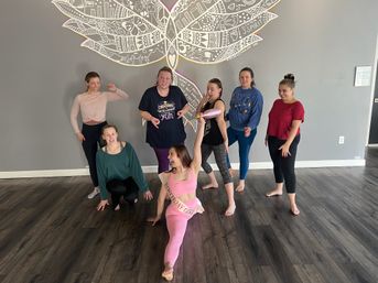 Group of seven women in an indoor yoga/fitness studio posing on dark wood floors in front of a large decorative mural; birthday woman in pink does a split wearing a sash while others smile and strike playful poses.