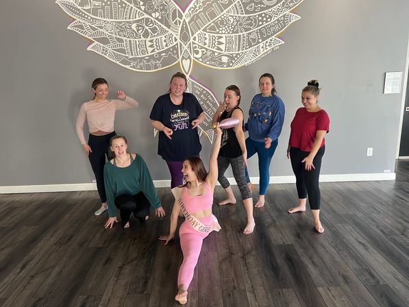 Group of seven women in an indoor yoga/fitness studio posing on dark wood floors in front of a large decorative mural; birthday woman in pink does a split wearing a sash while others smile and strike playful poses.