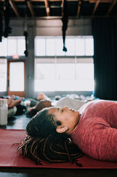 Person lying on a red yoga mat in savasana during a group relaxation class inside a sunlit indoor yoga studio with large windows