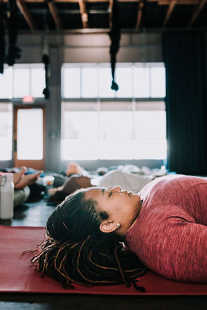Person lying on a red yoga mat in savasana during a group relaxation class inside a sunlit indoor yoga studio with large windows