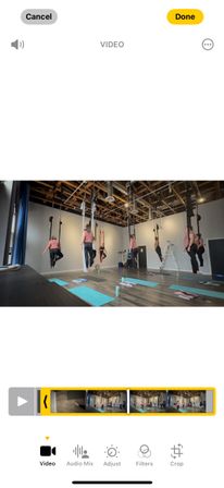 Screenshot of a phone video editor showing an indoor aerial yoga class — participants in pink tops suspended from ceiling silks above yoga mats in a modern studio with exposed wooden beams and workout gear.
