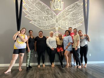Group of nine adults posing barefoot in an aerial yoga studio, smiling in front of a large decorative white butterfly mural on a gray wall with hanging aerial silks.