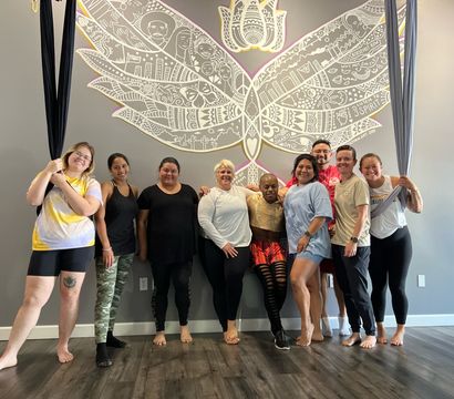 Group of nine adults posing barefoot in an aerial yoga studio, smiling in front of a large decorative white butterfly mural on a gray wall with hanging aerial silks.