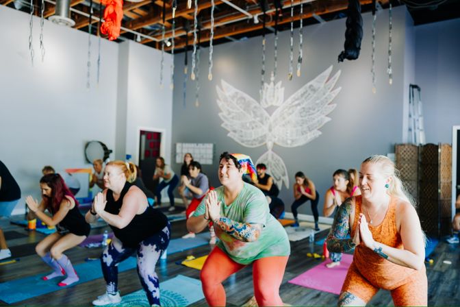 Energetic group yoga class in a bright indoor studio — diverse participants in colorful activewear holding chair pose with hands in prayer on mats beneath hanging aerial silks and a white wing mural