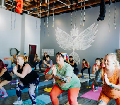 Energetic group yoga class in a bright indoor studio — diverse participants in colorful activewear holding chair pose with hands in prayer on mats beneath hanging aerial silks and a white wing mural