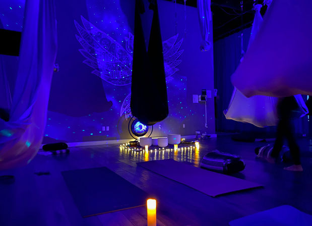 Blue-lit aerial yoga and meditation studio interior with hanging hammocks, floor mats, glowing candles, crystal singing bowls, and angel-wing mural
