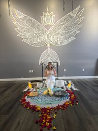 Woman kneeling in prayer pose before a round mandala altar with crystal singing bowls, candles and rose petals beneath a large white winged-lotus mural in a modern yoga and sound-bath studio.