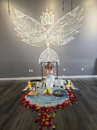 Woman kneeling in prayer pose before a round mandala altar with crystal singing bowls, candles and rose petals beneath a large white winged-lotus mural in a modern yoga and sound-bath studio.