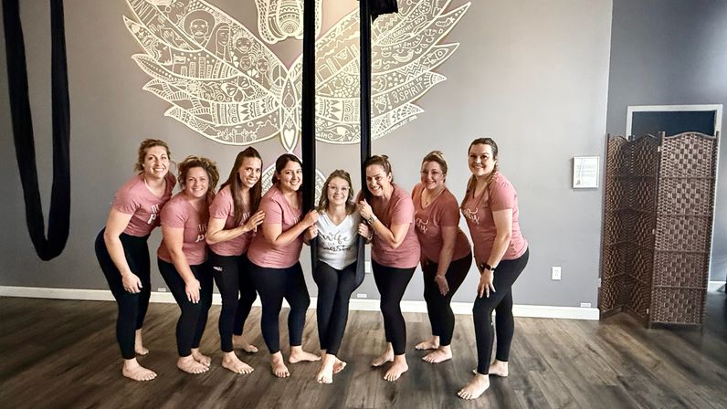 Nine women in matching pink shirts and black leggings posing barefoot around aerial silks in a yoga studio, a woman in a white top at center, with a large decorative wing mural on the gray wall behind them.