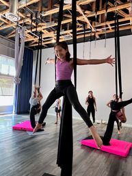 Smiling girl in a purple top performs a star-split on black aerial silks above bright pink crash mats in a sunlit indoor aerial-silks studio with other students.