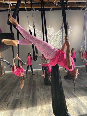 Children in pink shirts doing aerial-silks flips in an indoor aerial yoga studio, suspended on black fabric in front of a decorative wing mural over wood flooring.