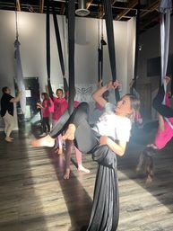 Kids aerial silks class in a sunlit dance studio, girls practicing hammock and inversion poses on black and white aerial silks suspended from the ceiling over wood floors