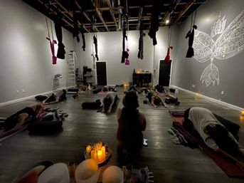 Dim, cozy aerial yoga studio interior with suspended hammocks from exposed beams, students on mats in restorative child's pose around glowing candles and a lotus wall mural.