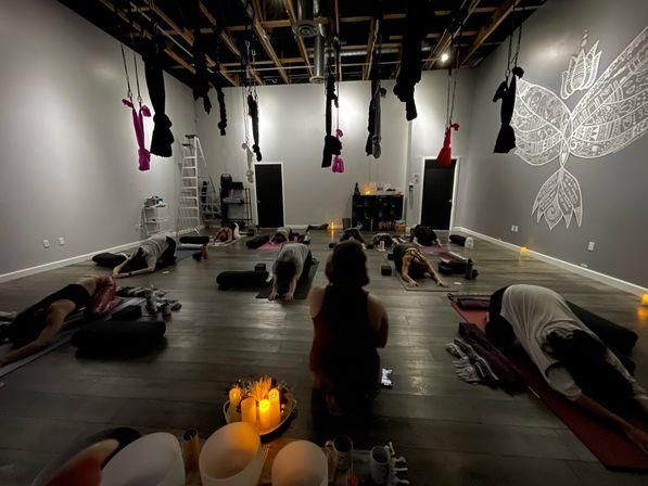 Dim, cozy aerial yoga studio interior with suspended hammocks from exposed beams, students on mats in restorative child's pose around glowing candles and a lotus wall mural.