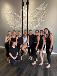 Aerial yoga class group photo: nine women in activewear gathered around black aerial silks, barefoot and smiling in front of a decorative white wing mural on a gray studio wall.