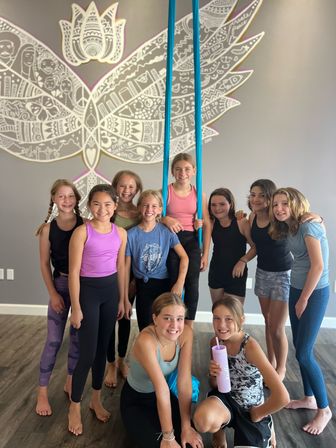 Smiling girls in activewear posing in an indoor aerial-silks studio, one on bright blue silks, in front of a large white wing-and-lotus mural on a gray wall and wood floor.