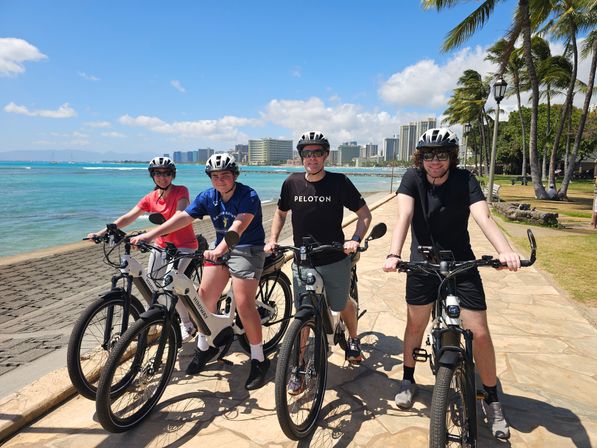 Four helmeted cyclists on electric bikes paused on a sunny oceanfront promenade with palm trees, turquoise water and a tropical city skyline in the background.