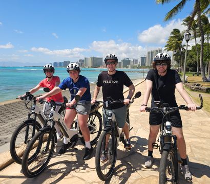 Four helmeted cyclists on electric bikes paused on a sunny oceanfront promenade with palm trees, turquoise water and a tropical city skyline in the background.