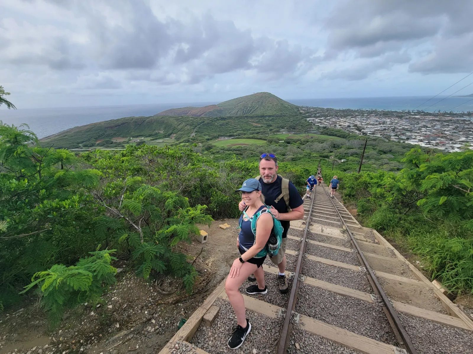 Couple hiking up steep disused railroad tracks on a lush tropical coastal ridge, scenic ocean and town below under a dramatic cloudy sky — adventurous coastal trail view.