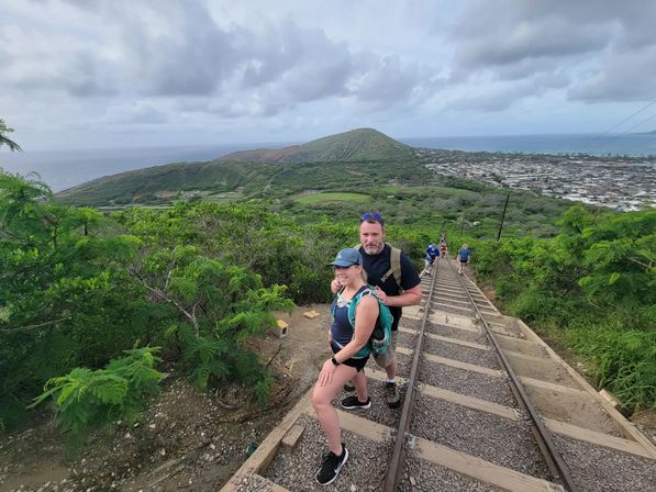 Couple hiking up steep disused railroad tracks on a lush tropical coastal ridge, scenic ocean and town below under a dramatic cloudy sky — adventurous coastal trail view.