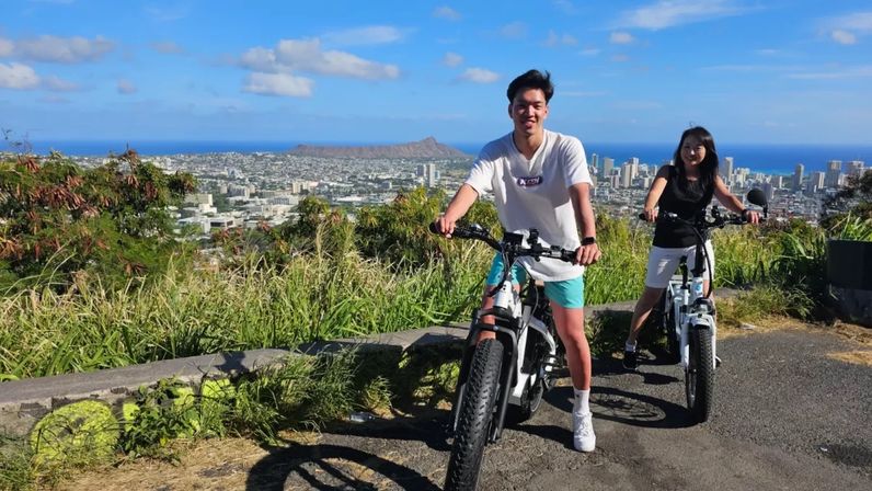 Two riders on electric bikes at a grassy hillside overlook above Honolulu, with Waikiki skyline and Diamond Head crater framed by the blue Pacific and sunny sky.