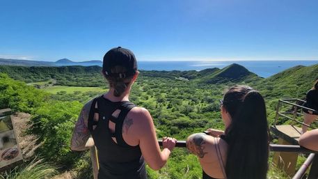 Two hikers at a lookout railing admiring a panoramic tropical coastal view of a lush volcanic crater, rolling green hills and the sparkling Pacific Ocean under a clear blue sky.
