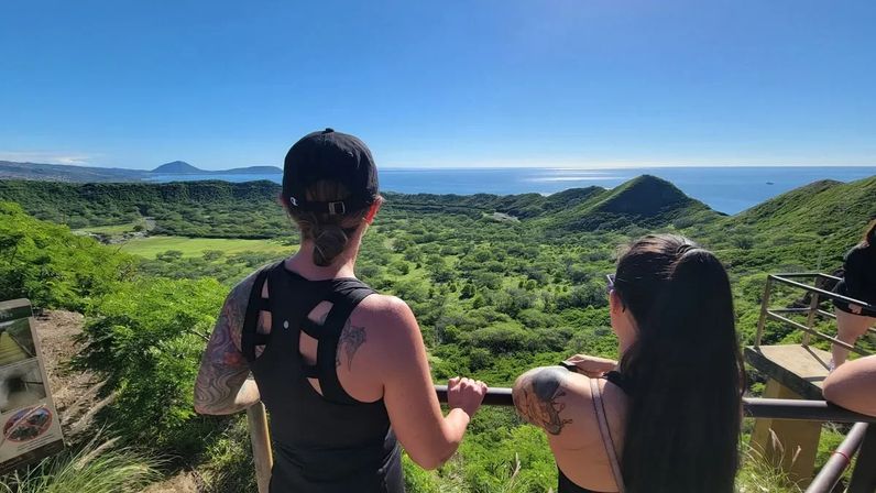 Two hikers at a lookout railing admiring a panoramic tropical coastal view of a lush volcanic crater, rolling green hills and the sparkling Pacific Ocean under a clear blue sky.