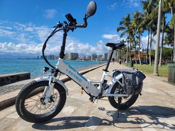 White electric folding bike with rear pannier parked on a sunny palm‑lined seaside promenade, turquoise ocean and distant city skyline under a bright blue sky, ready for a waterfront ride.