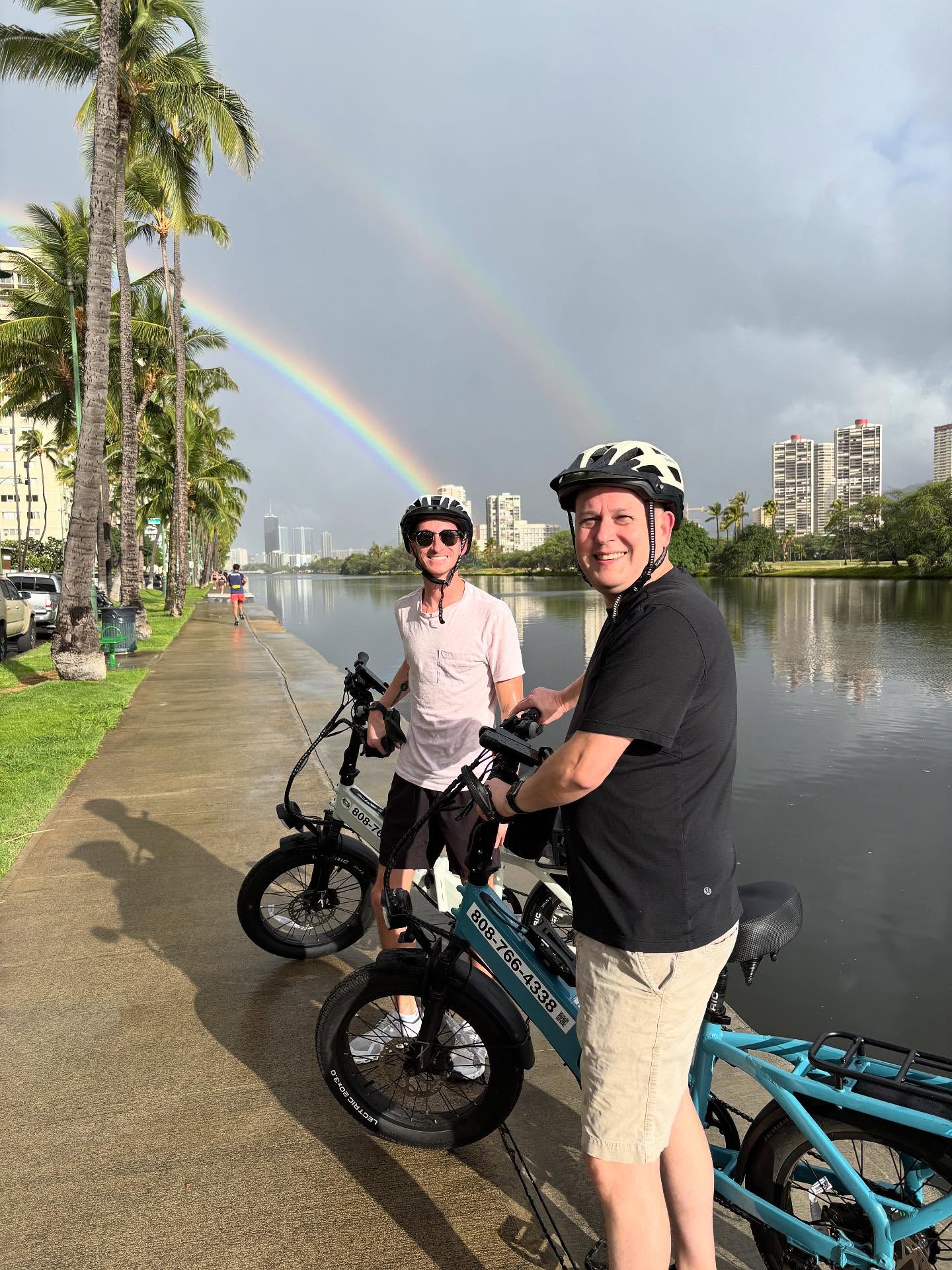 Two grinning cyclists in helmets on electric bikes along a palm-lined urban canal and city skyline beneath a vivid double rainbow after a rain shower