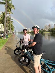 Two grinning cyclists in helmets on electric bikes along a palm-lined urban canal and city skyline beneath a vivid double rainbow after a rain shower