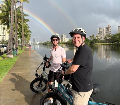 Two grinning cyclists in helmets on electric bikes along a palm-lined urban canal and city skyline beneath a vivid double rainbow after a rain shower