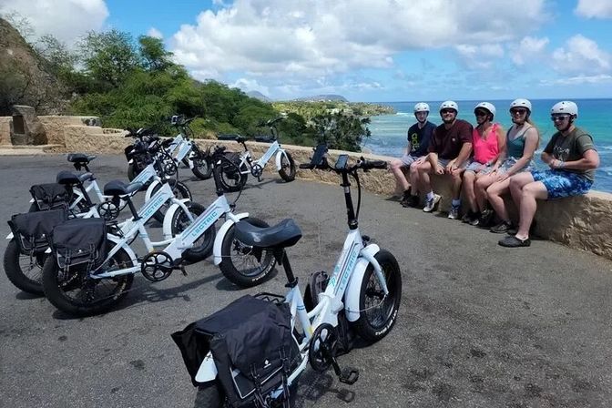E-bike tour at a tropical coastal overlook — white electric bikes parked on pavement, five helmeted riders sitting on a stone wall with turquoise ocean, sandy beach and green cliffs under a blue sky.